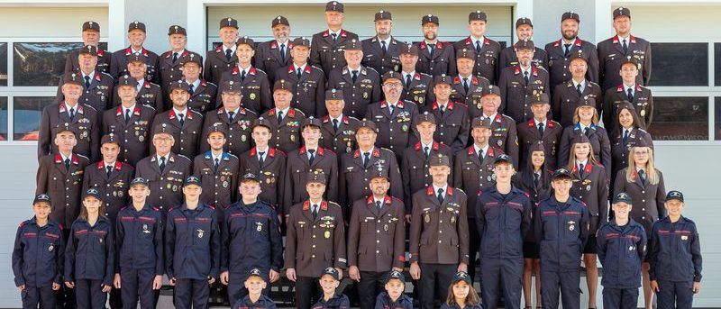 Eine Gruppenaufnahme von uniformierten Personen, einschließlich Männer und Frauen, die in Reihen stehen, mit einem Gebäude im Hintergrund. Die Gruppe besteht aus jungen Menschen, Erwachsenen und einer Mischung aus Geschlechtern. Sie tragen Hüte, Krawatten und Medaillen, wobei einige Personen eine Brille tragen. Sie alle lächeln und posieren für das Foto.