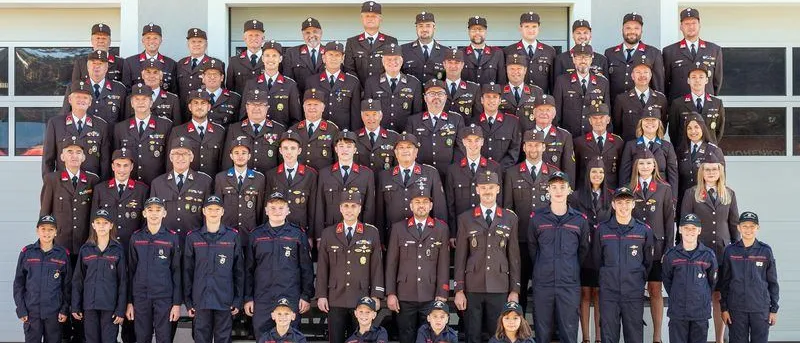 Eine Gruppenaufnahme von uniformierten Personen, einschließlich Männer und Frauen, die in Reihen stehen, mit einem Gebäude im Hintergrund. Die Gruppe besteht aus jungen Menschen, Erwachsenen und einer Mischung aus Geschlechtern. Sie tragen Hüte, Krawatten und Medaillen, wobei einige Personen eine Brille tragen. Sie alle lächeln und posieren für das Foto.