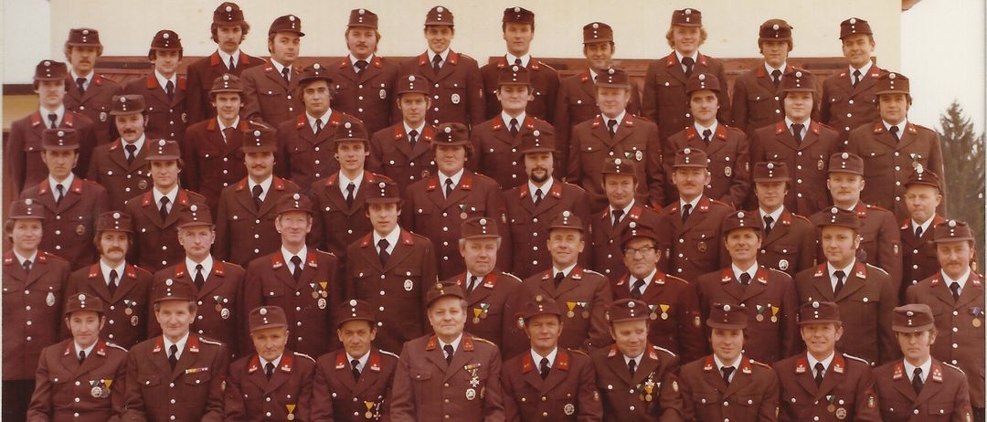 Ein Gruppenfoto von Feuerwehrleuten in traditionellen Uniformen, die vor einem Gebäude mit dem Namen Freiwillige Feuerwehr Hohenkogl-Mitterdorf posieren. Sie tragen alle Mützen, Krawatten und Abzeichen, einige haben Medaillen.