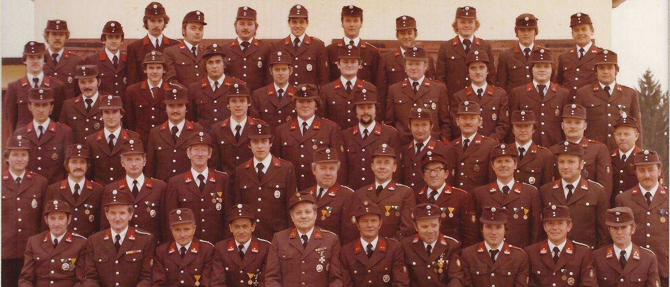 Ein Gruppenfoto von Feuerwehrleuten in traditionellen Uniformen, die vor einem Gebäude mit dem Namen Freiwillige Feuerwehr Hohenkogl-Mitterdorf posieren. Sie tragen alle Mützen, Krawatten und Abzeichen, einige haben Medaillen.