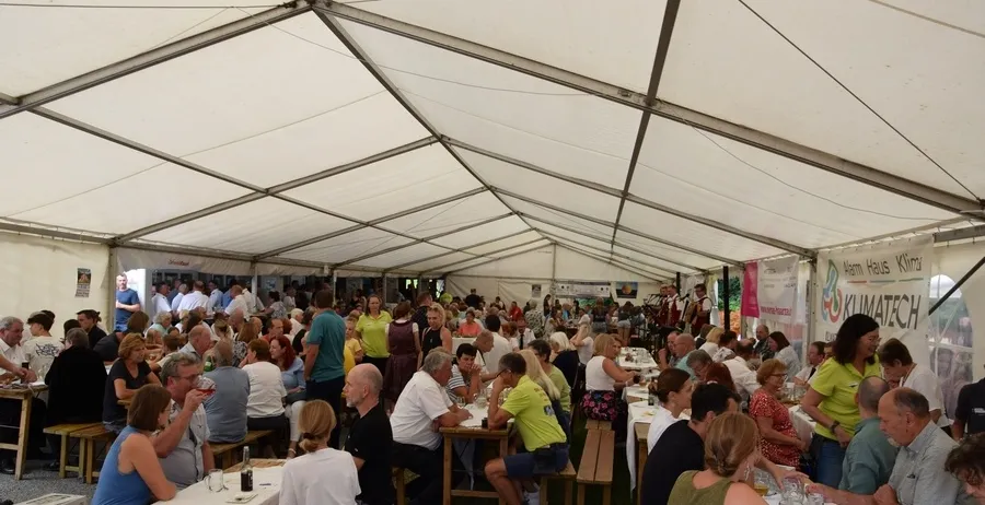 Viele Menschen sind zu einer Veranstaltung im Freien unter einem großen Zelt versammelt, sitzen an Tischen und essen. Einige tragen Brillen, Hüte und Turnschuhe. Der Bereich ist mit grünem Gras bedeckt.