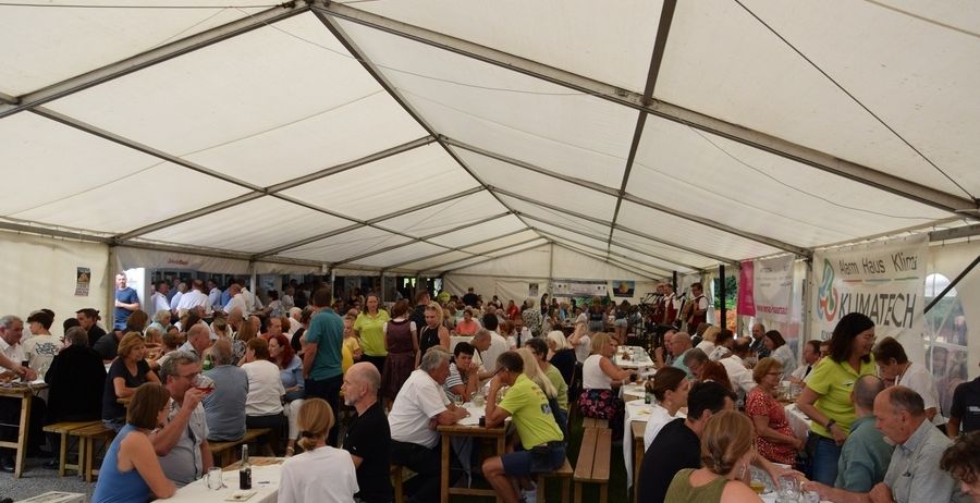 Viele Menschen sind zu einer Veranstaltung im Freien unter einem großen Zelt versammelt, sitzen an Tischen und essen. Einige tragen Brillen, Hüte und Turnschuhe. Der Bereich ist mit grünem Gras bedeckt.