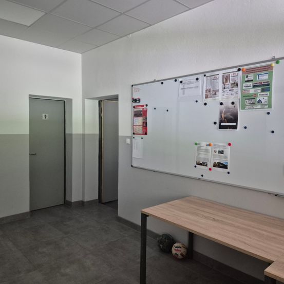 Ein leerer Raum mit einer weißen Pinnwand an der Wand, einem Holztisch und einer geschlossenen Tür.