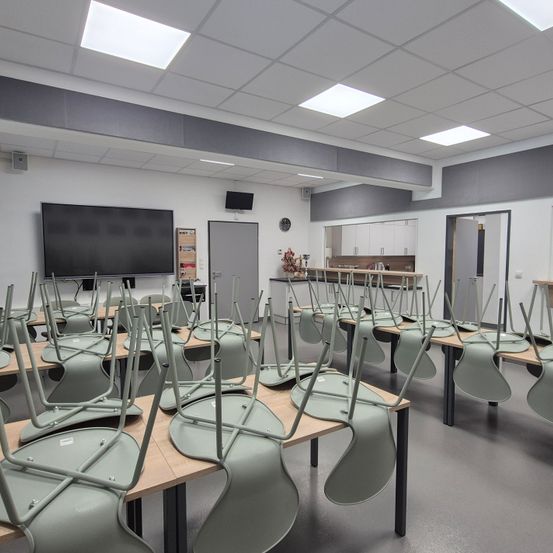 Ein leerer Klassenraum mit mehreren Tischen und Stühlen, einem großen Flachbildfernseher an der Wand und einer Uhr über der Tür.