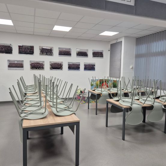 Ein leerer Klassenraum mit grauen Stühlen in Reihen angeordnet. Die Wände sind mit Fotos von Gruppen von Menschen dekoriert. Der Raum hat eine Decke mit quadratischen Lichtern.