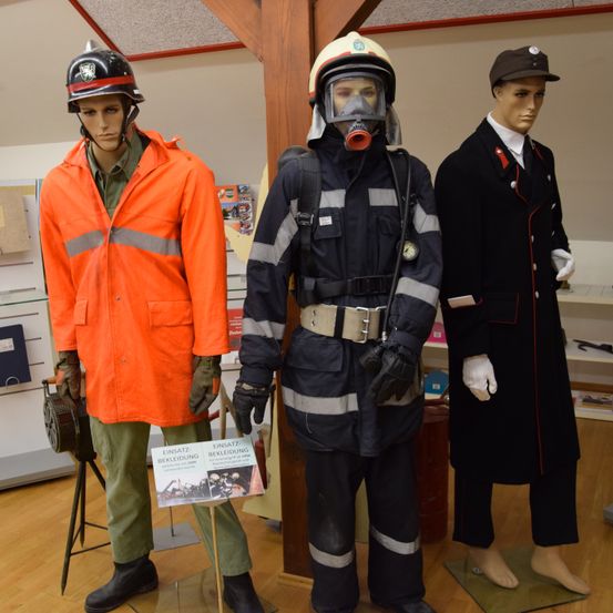 Drei Mannequins in verschiedenen Uniformen und Helmen sind in einem Raum ausgestellt, möglicherweise in einem Museum. Ein Schild steht vor dem mittleren Mannequin.