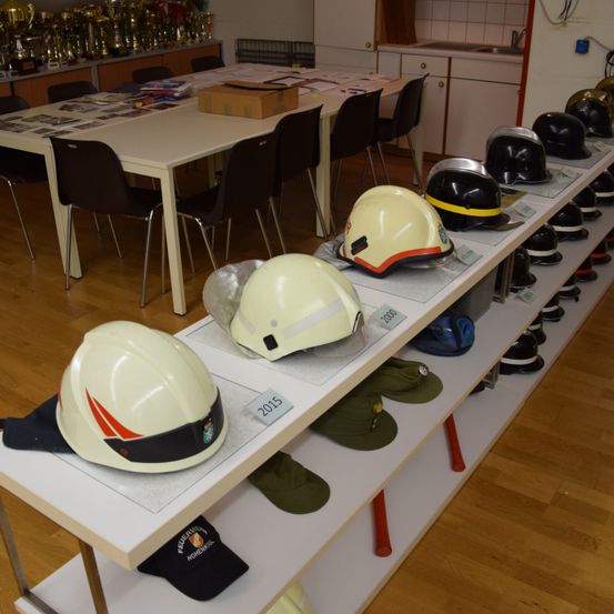 Eine Reihe von Helmen, die auf einem Regal ausgestellt sind, mit mehreren Helmen, die mit Jahren wie 2000, 2015 und anderen beschriftet sind. Im Hintergrund befindet sich ein langer Tisch mit Stühlen.