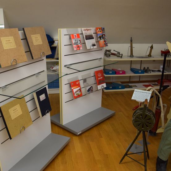 Eine Innenausstellung von Büchern und Dokumenten auf Glasregalen. Ein Holzboden und eine weiße Wand sind sichtbar. Ein Schild auf einem Stativ steht 'Krukkenspritze'.