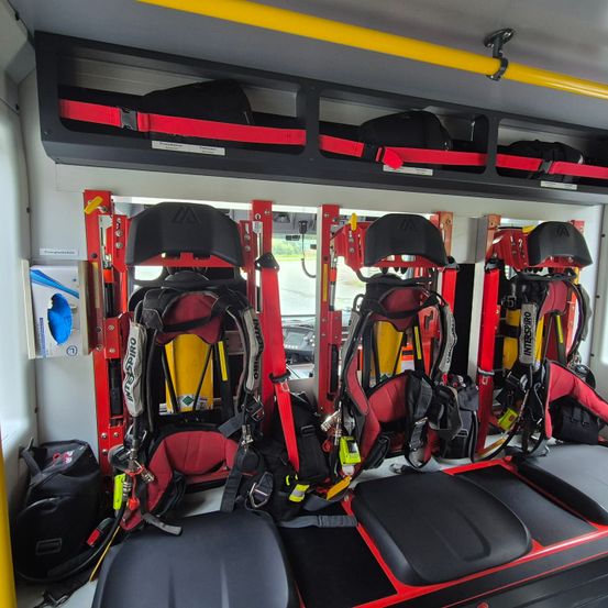 Das Innere eines Rettungswagens, ausgestattet mit Sicherheitsgurten und medizinischer Ausrüstung. Vier rote und schwarze Sitze sind an roten Rahmen montiert. Taschen und Ausrüstung sind auf den Regalen aufbewahrt.