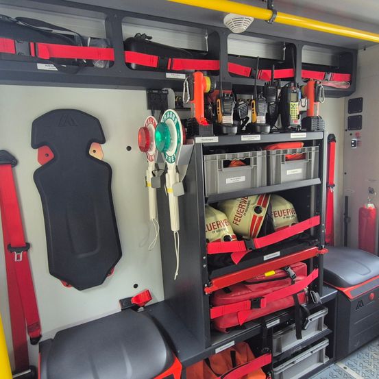 Innenraum eines Rettungsfahrzeugs, zeigt organisierte Fächer mit beschrifteten Aufbewahrungsboxen, Feuerlöschern, Helmen, Funkgeräten und diversen Werkzeugen. Rote und gelbe Gurte sichern Gegenstände.
