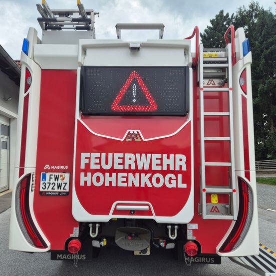 Die Rückseite eines roten und weißen Feuerwehrfahrzeuges mit einem leuchtenden Warnzeichen auf der Rückseite. Das Schild steht für 'FEUERWEHR HOHENKOGL'. Das Nummernschild des Fahrzeugs lautet 'FW 372WZ'.