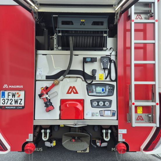 Die Rückseite eines roten Feuerwehrwagens, gekennzeichnet mit FW 372WZ, zeigt verschiedene Anzeigen, ein Funkgerät und eine Leiter. An der Seite befinden sich ein roter Schlauchaufsatz und ein Feuerlöscher.