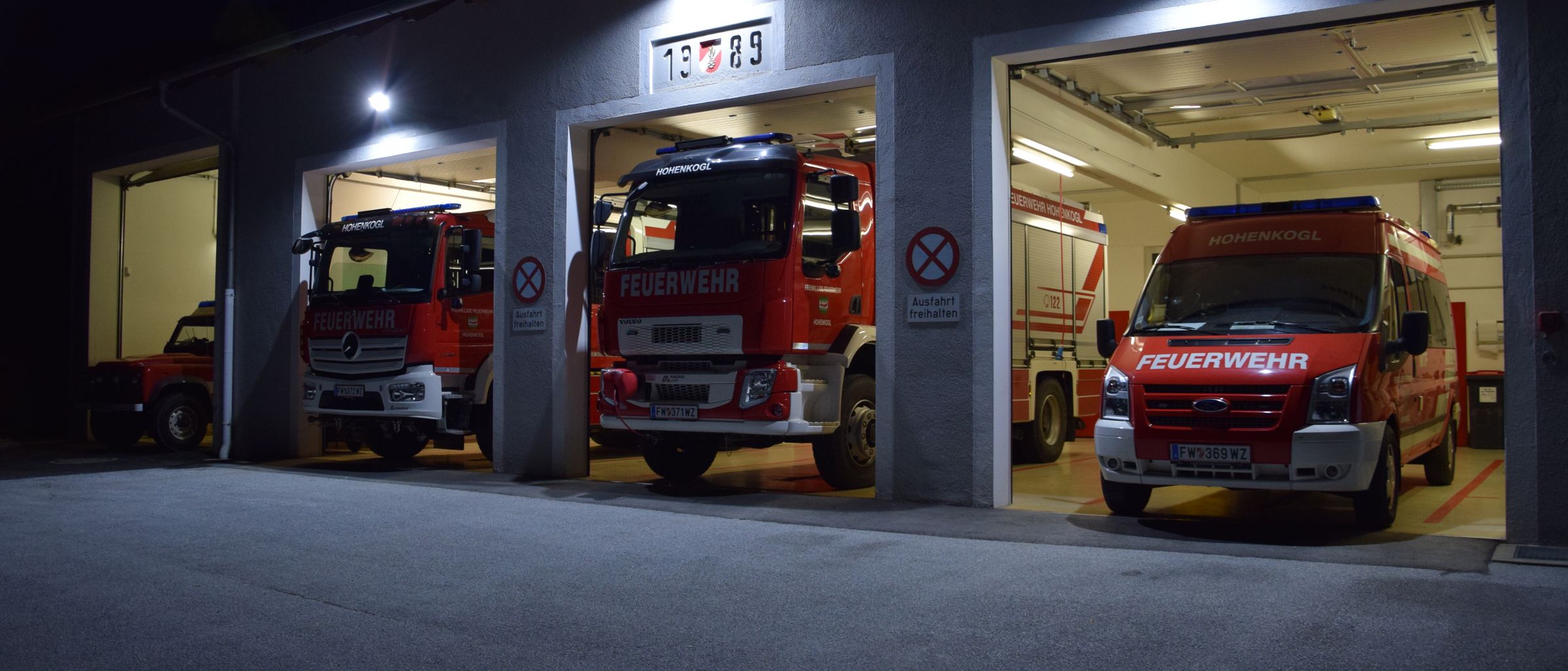 In der Nacht ist eine Feuerwache mit drei Notfallfahrzeugen im Inneren geparkt. Das mittlere Fahrzeug hat die Nummer 13.7.89. Es gibt Schilder, die Rauchen und Fahren verbieten.