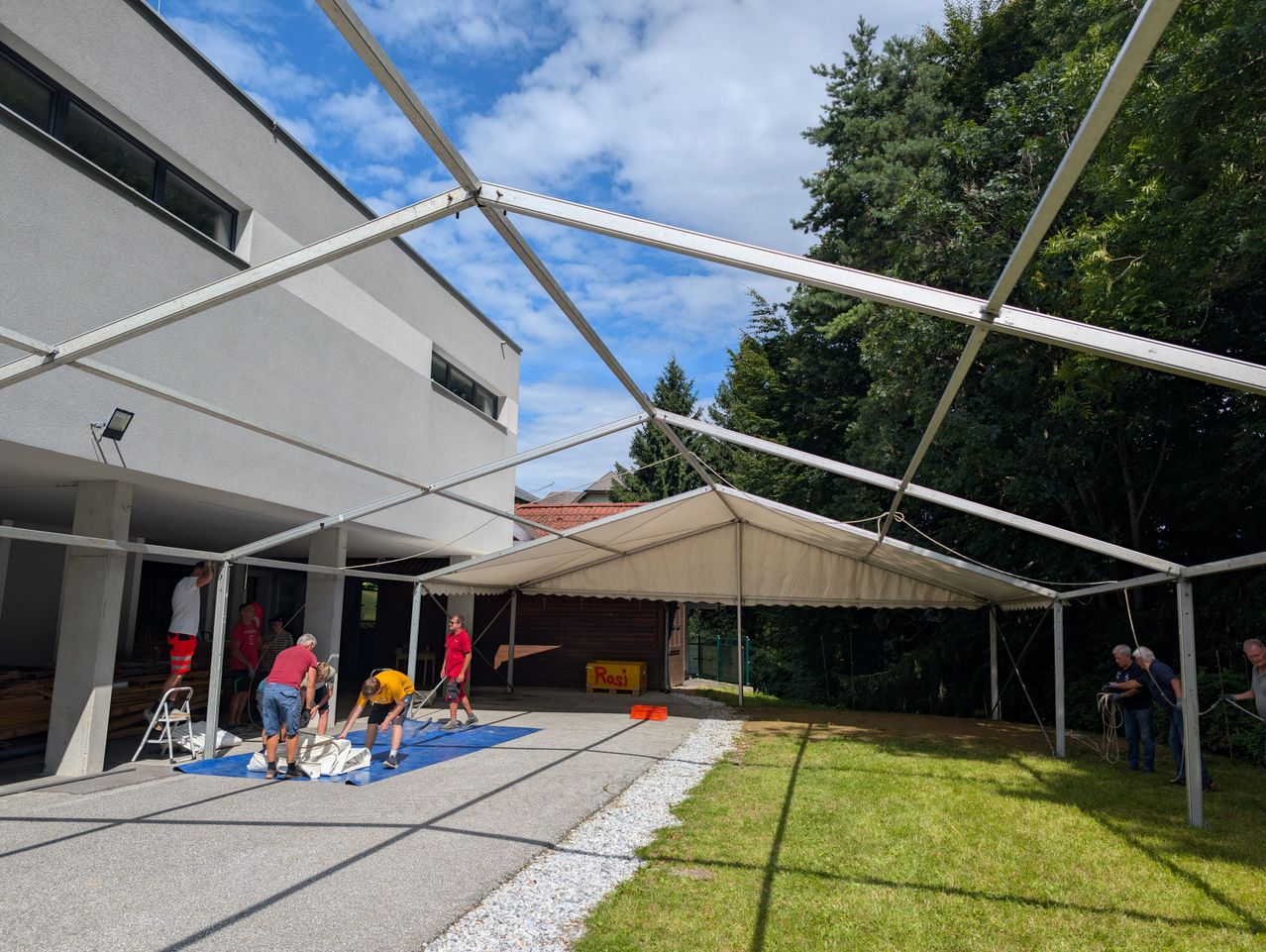 Eine Gruppe von Menschen richtet ein großes weißes Zelt vor einem Gebäude an einem sonnigen Tag auf.