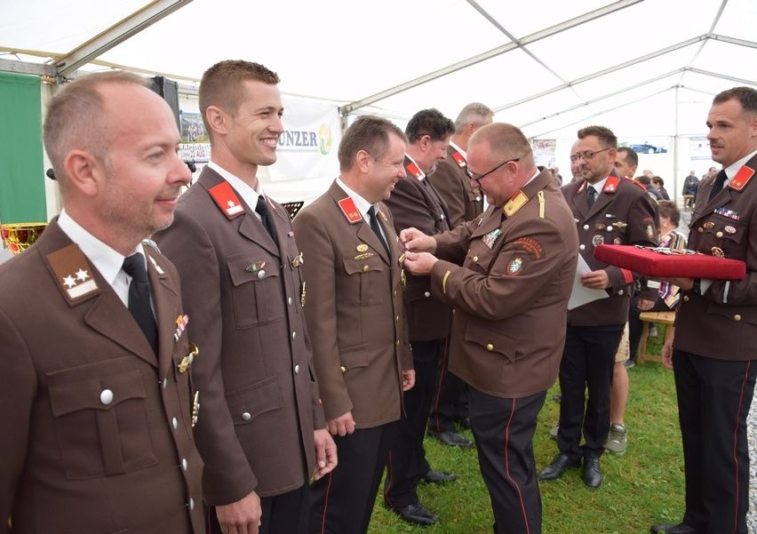 Eine Gruppe von Männern in Militäruniformen steht unter einem Zelt, wobei ein Offizier Medaillen auf die Brust anderer heftet. Sie lächeln und scheinen sich bei einer formellen Veranstaltung zu befinden.