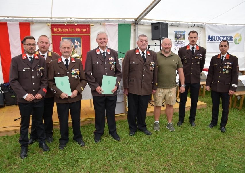 Sieben Männer stehen auf Gras und tragen Uniformen und halten Urkunden. Ein Banner hinter ihnen sagt 'Munzel' und es gibt italienische Flaggen.