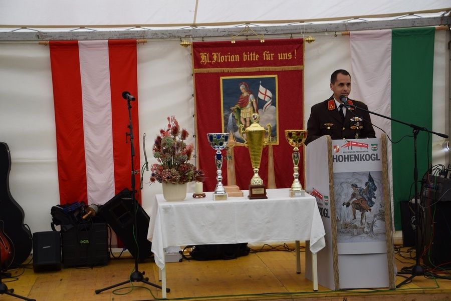 Ein Mann in Uniform steht an einem Podium mit Mikrofonen vor einem Tisch mit Trophäen und einer Fahne dahinter. Die Bühne ist unter einem Zelt mit Flaggen aufgebaut.