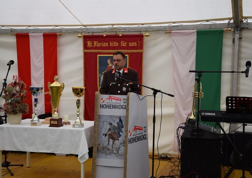 Ein Mann in Uniform steht an einem Podium mit Trophäen auf einem Tisch. Hinter ihm ist ein Banner und Flaggen. Rechts befindet sich ein Yamaha-Keyboard und eine Trompete.