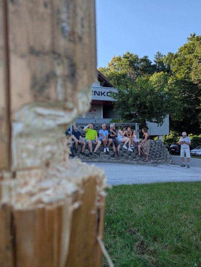 Eine Gruppe von Menschen sitzt auf einer Betonkante und beobachtet etwas. Sie sind in der Nähe eines Gebäudes mit ENKO-Beschilderung. Bäume und geparkte Autos sind im Hintergrund.