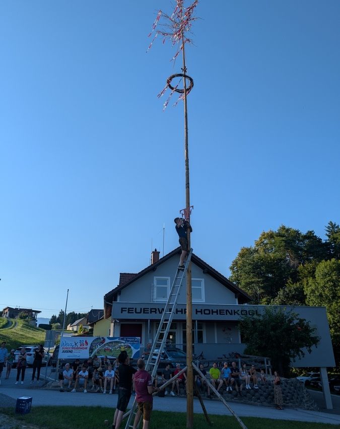 Ein Mann klettert eine Leiter hinauf zu einem mit Dekorationen geschmückten Pfahl, während die Menschen auf dem Dorfplatz zusehen. Der Himmel ist klar und es gibt ein Gebäude mit dem Schild Feuerwache Hohenkogel.