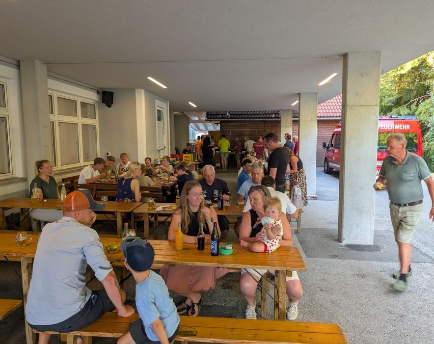 Eine Gruppe von Menschen diniert in einem Außenbereich mit Holztischen und Bänken. Flaschen, Gläser und Essen sind auf den Tischen. Ein Feuerwehrauto ist in der Nähe geparkt.