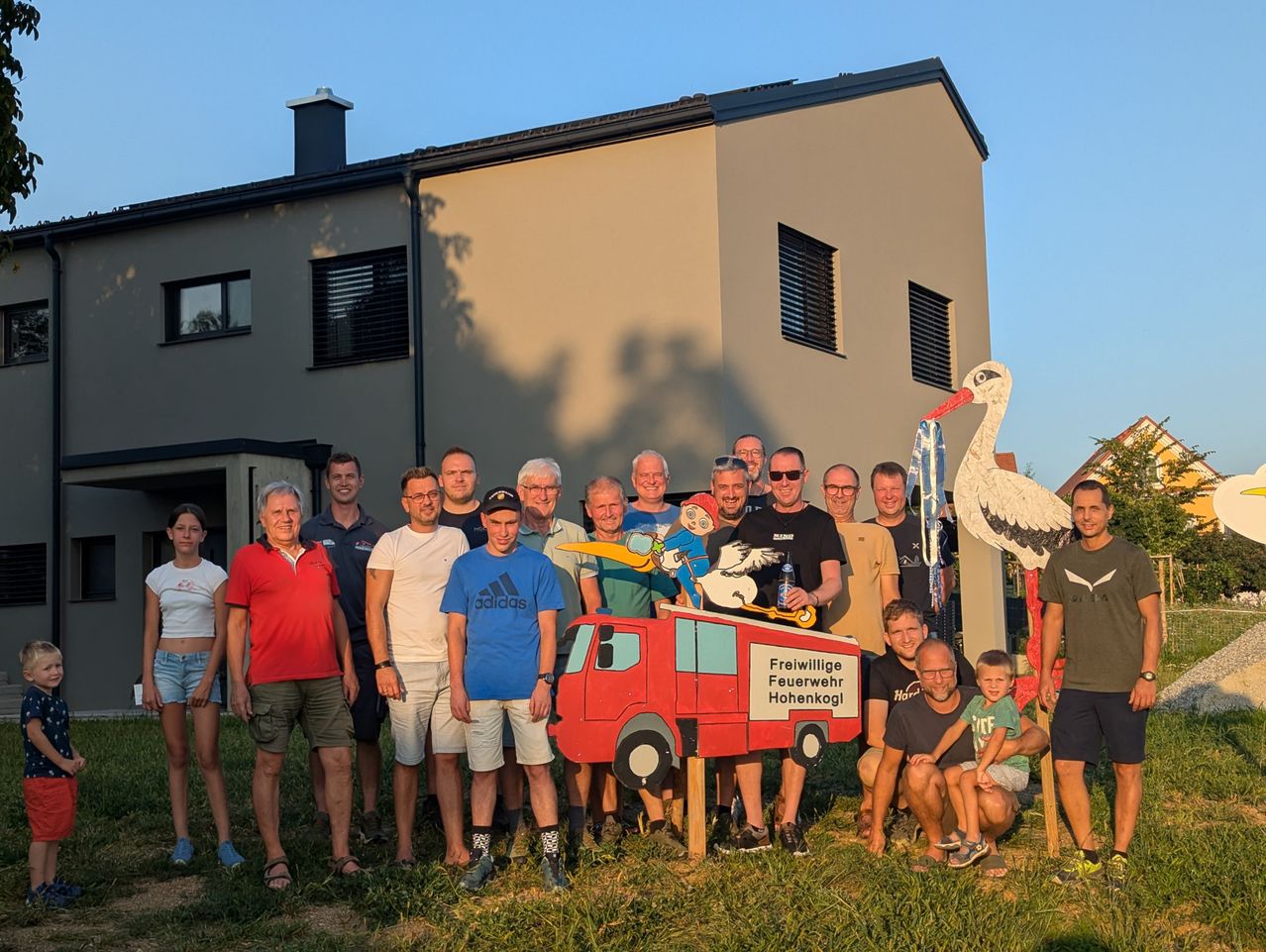 Eine Gruppe von Menschen steht vor einem modernen Haus. Sie lächeln und scheinen für ein Foto zu posieren. Ein rotes Feuerwehrauto-Modell steht in der Mitte, und eine Storchstatue ist rechts. Der Himmel ist klar.