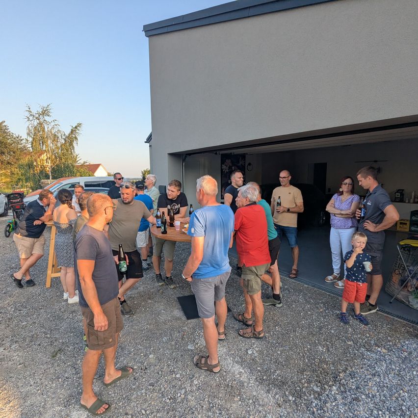 Eine Gruppe von Menschen versammelt sich vor einem Haus, einige sitzen um einen Tisch mit Getränken, während andere stehen und sich unterhalten. Die Umgebung ist im Freien mit Kiesboden und Bäumen im Hintergrund.