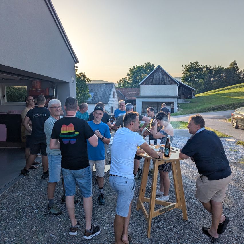 Eine Gruppe von Menschen steht und unterhält sich um einen Holztisch. Sie haben Flaschen und Gläser auf dem Tisch. Einige tragen Brillen, Turnschuhe und Mützen. Dahinter steht ein Gebäude mit einer Garage. In der Ferne sind Häuser, ein Auto und Bäume.