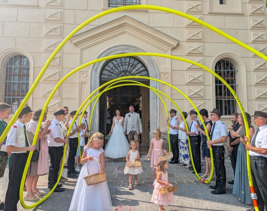Eine Hochzeitsgesellschaft geht durch einen Bogen aus gelben Rohren. Die Braut und der Bräutigam stehen in der Mitte, umgeben von Kindern mit Körben. Sie stehen vor einem Gebäude mit einem dekorierten Bogen.