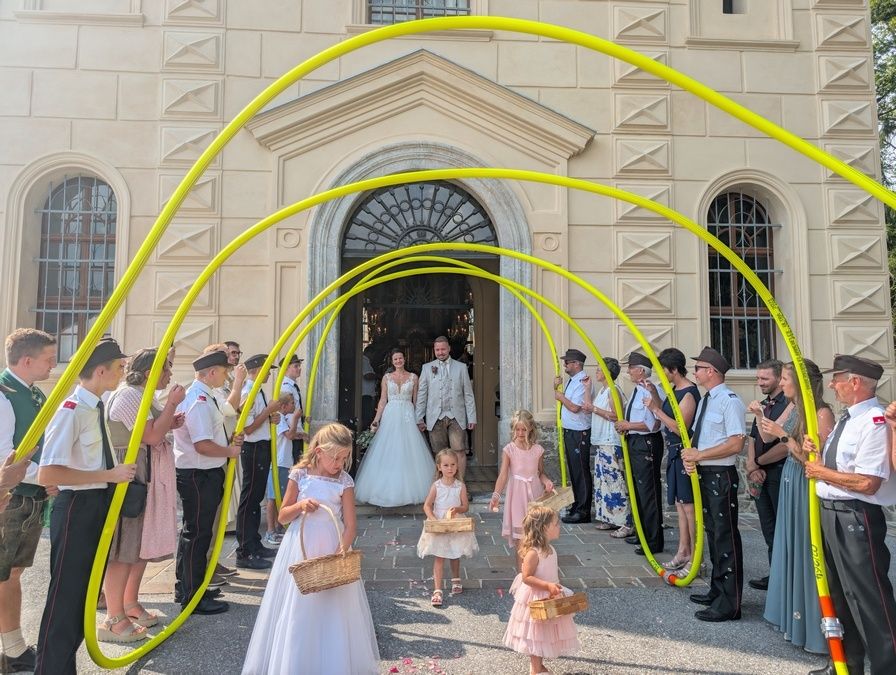 Eine Hochzeitsgesellschaft geht durch einen Bogen aus gelben Rohren. Die Braut und der Bräutigam stehen in der Mitte, umgeben von Kindern mit Körben. Sie stehen vor einem Gebäude mit einem dekorierten Bogen.
