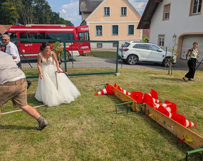 Eine Braut in einem Hochzeitskleid spielt mit einer Gruppe von Menschen ein Spiel auf einem Rasen. Im Hintergrund steht ein Feuerwehrauto und ein Haus.