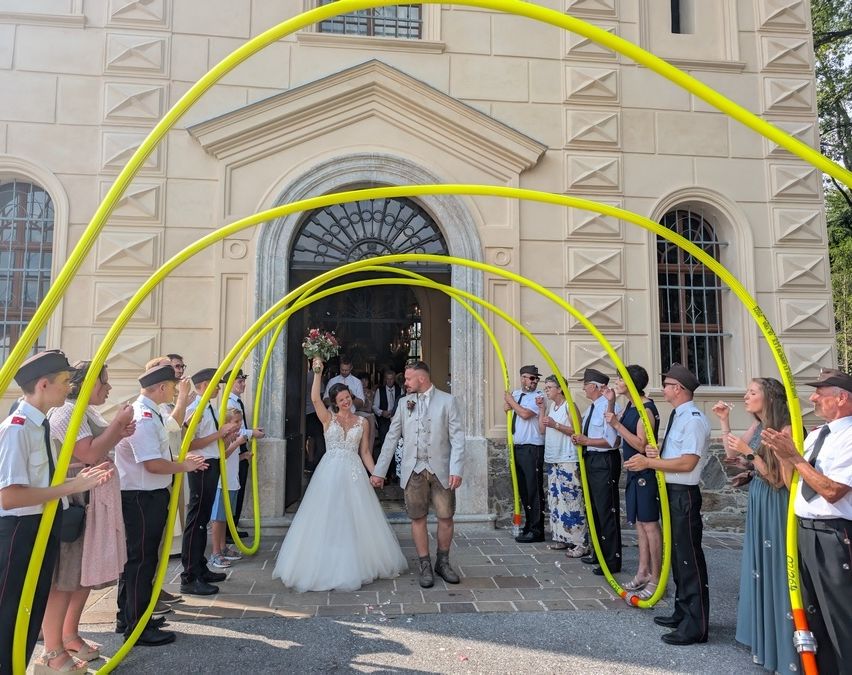 Ein frisch verheiratetes Paar geht durch einen gelben Bogen bei einer Hochzeit, umgeben von Zuschauern. Die Braut hält einen Blumenstrauß, und der Bräutigam trägt einen weißen Anzug. Sie stehen vor einem Gebäude mit einer dekorativen Fassade.