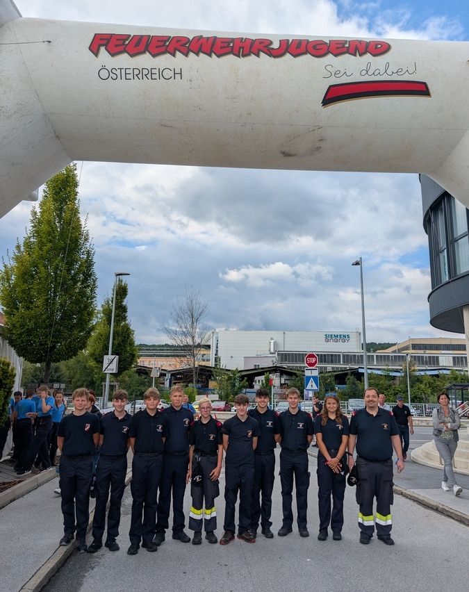 Eine Gruppe junger Feuerwehrleute steht unter einem Banner mit der Aufschrift 'Feuerwehrjugend Osterreich', mit einem Gebäude und einer Straße im Hintergrund.