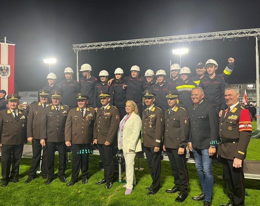 Eine Gruppe uniformierter Personen steht auf einem Rasenfeld, einige tragen Helme. Dahinter befindet sich eine Metallstruktur mit Lichtern. Sie posieren für ein Foto bei Nacht.