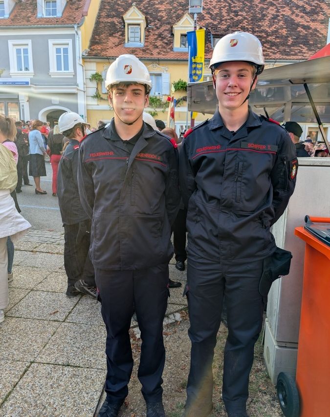 Zwei junge Männer in Feuerwehruniformen stehen vor einem Gebäude mit einer Menschenmenge im Hintergrund. Sie tragen beide weiße Helme.