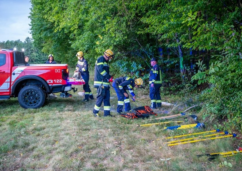 Feuerwehrleute sind neben einem roten Pickup-Truck im Freien und arbeiten im Gras an etwas. Sie tragen Helme und Schutzausrüstung. In der Nähe liegen Werkzeuge und Ausrüstung auf dem Boden.