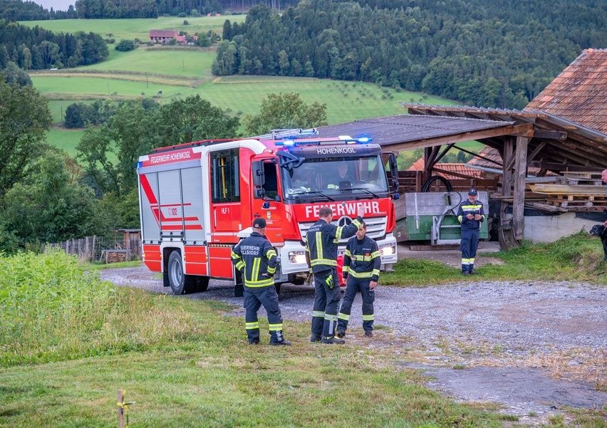 Ein Feuerwehrauto steht auf einer Kiesauffahrt. Vier Feuerwehrleute stehen neben dem Fahrzeug, einer davon auf der Seite. In der Nähe befindet sich ein Schuppen mit Dach.