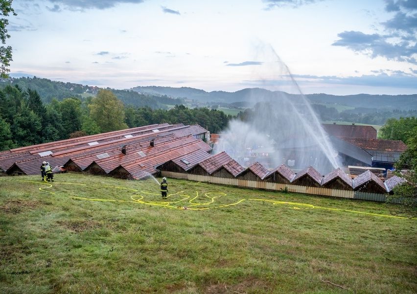 Feuerwehrleute sprühen Wasser auf ein Dach mit Solarpanelen. Zwei Personen sind auf dem Dach und zwei am Boden. Die Landschaft ist ländlich mit Hügeln und Bäumen.