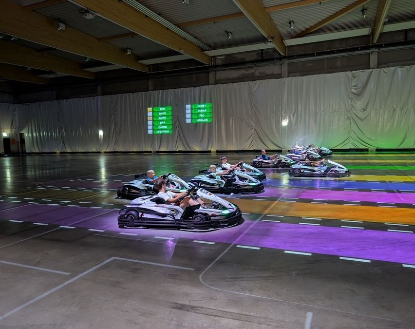 Eine Gruppe von Menschen fährt Go-Karts auf einer farbenfrohen Rennstrecke in einem großen Innenbereich mit einer digitalen Anzeigetafel.