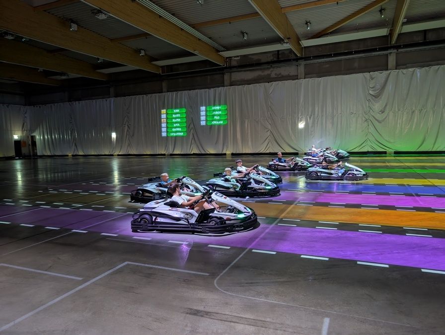 Eine Gruppe von Menschen fährt Go-Karts auf einer farbenfrohen Rennstrecke in einem großen Innenbereich mit einer digitalen Anzeigetafel.
