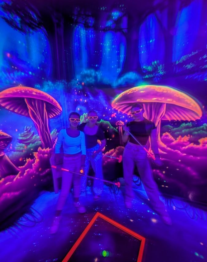 Drei Personen in einem neonbeleuchteten Indoor-Wald, mit Sonnenbrillen und Golfschlägern, posieren vor leuchtenden Pilzen.