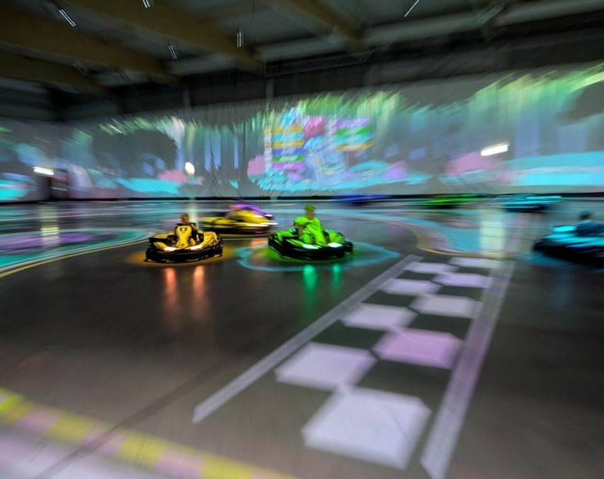In einer Go-Kart-Rennanlage fahren zwei Rennfahrer auf einer Strecke mit Neonlichtern. Die Strecke hat Schachbrettmuster auf dem Boden. Die Wände sind mit farbigen Projektionen beleuchtet.
