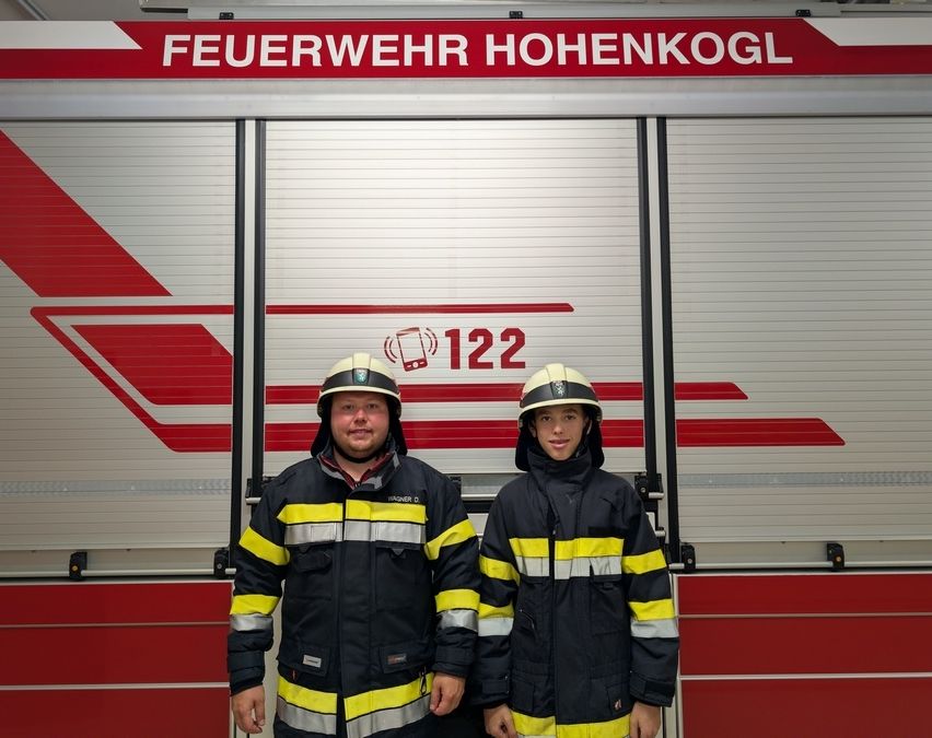 Zwei Feuerwehrleute stehen vor einem Feuerwehrwagen mit der Aufschrift 'Feuerwehr Hohenkogl 122'. Sie tragen identische Uniformen mit reflektierenden Streifen.