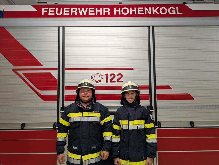 Zwei Feuerwehrleute stehen vor einem Feuerwehrwagen mit der Aufschrift 'Feuerwehr Hohenkogl 122'. Sie tragen identische Uniformen mit reflektierenden Streifen.