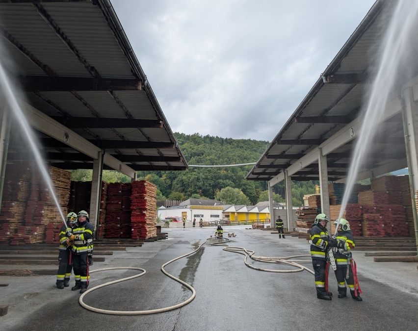Feuerwehrleute üben in einem Lagerhaus, indem sie Wasser aus Schläuchen sprühen. Stapel von Holz umgeben sie, und es gibt Gebäude im Hintergrund.
