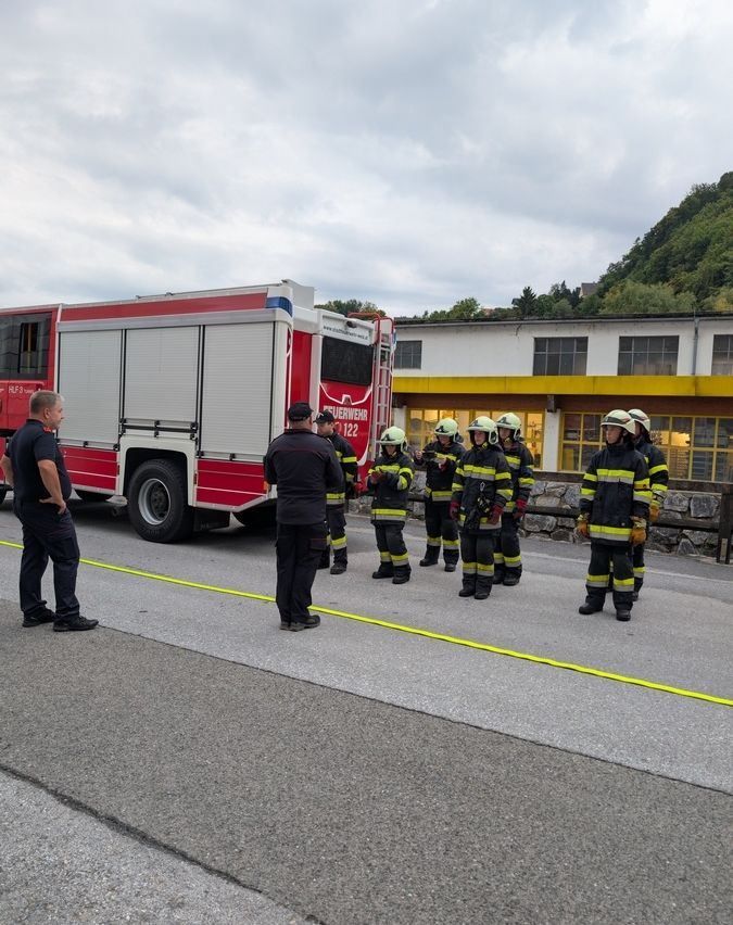 Eine Gruppe Feuerwehrleute in voller Ausrüstung steht vor einem Feuerwehrwagen, mit zwei Männern in Uniformen in der Nähe. Die Szene spielt vor einem Gebäude mit gelben Akzenten.