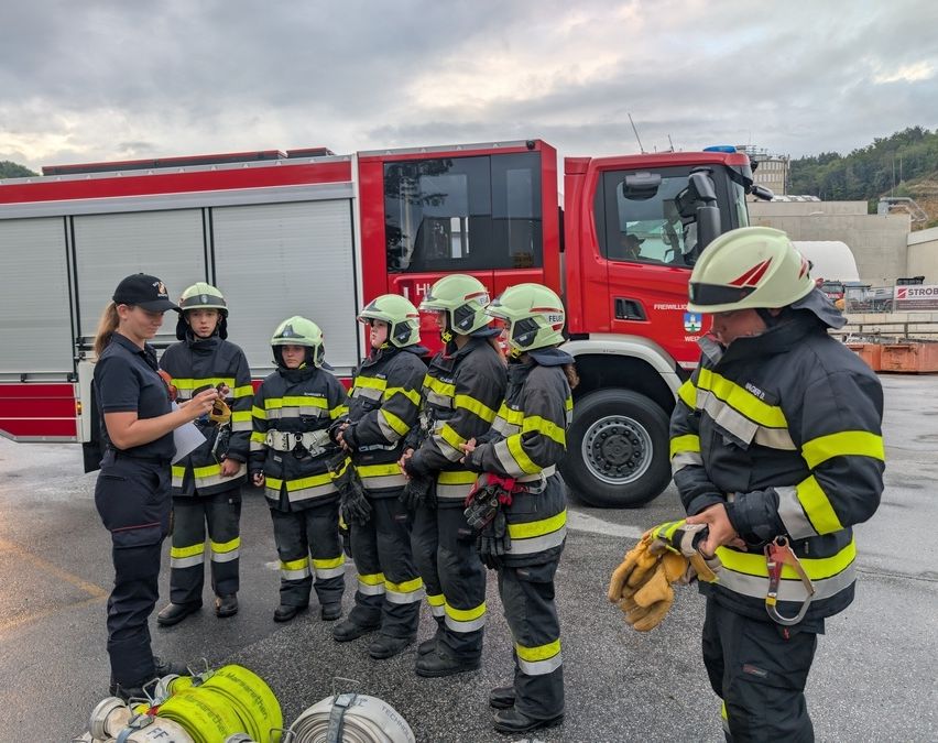 Eine Gruppe von Feuerwehrleuten in Uniformen versammelt sich auf einem Parkplatz, wahrscheinlich zum Training. Eine Frau vor ihnen hält ein Papier. Vor ihnen steht ein roter Feuerwehrwagen.