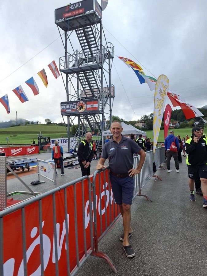 Ein Mann lehnt sich an ein Metallgeländer mit einem Banner dahinter. Flaggen hängen von einem Turm im Hintergrund. Andere Leute gehen auf der Straße.