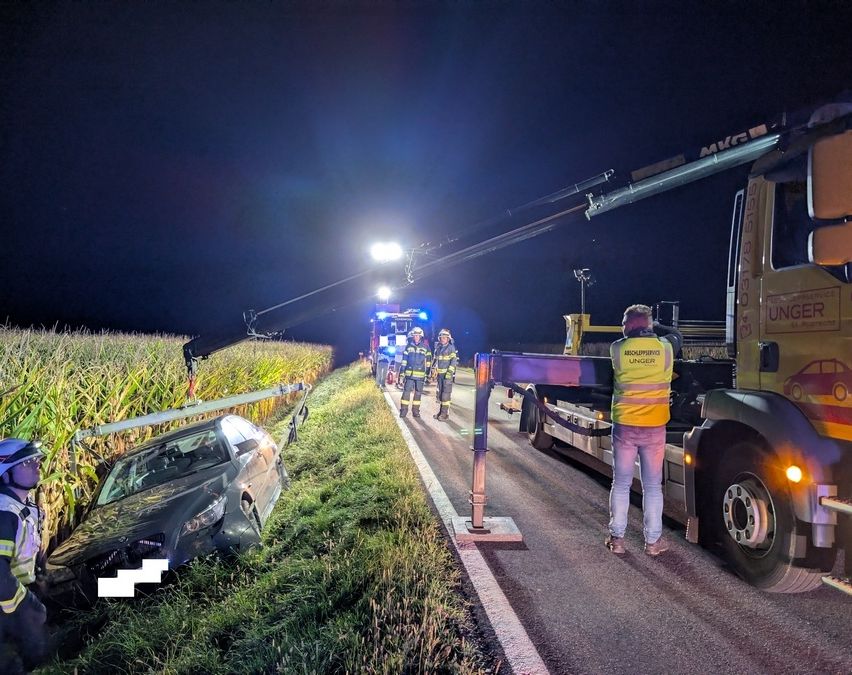 In der Nacht ein Unfallort auf einer Landstraße. Ein Abschleppwagen hebt ein beschädigtes Auto, und Rettungskräfte sind in der Nähe.