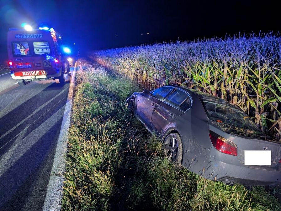 Ein Krankenwagen mit blinkenden Lichtern steht neben einem beschädigten Auto am Straßenrand. Das Auto ist im Grasfeld gekippt.